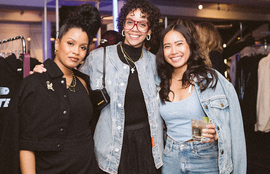 Three women enjoying a cocktail with racks of clothing behind
